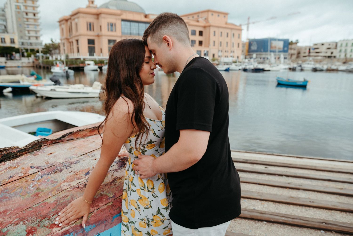Intimate moment between couple on wooden dock with boats and pink building in marina background.