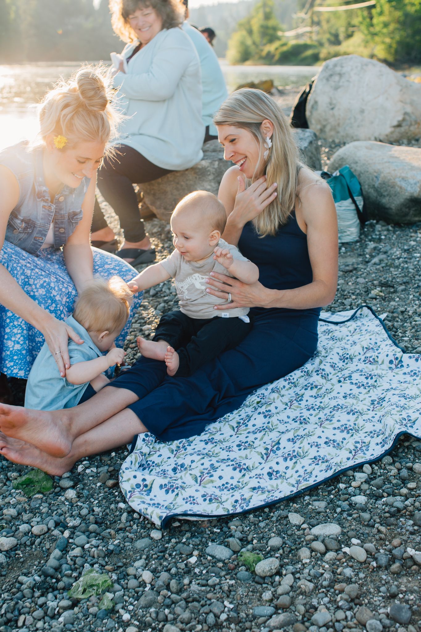 Two mothers sit on a rocky beach holding babies in golden sunlight, smiling warmly outdoors.