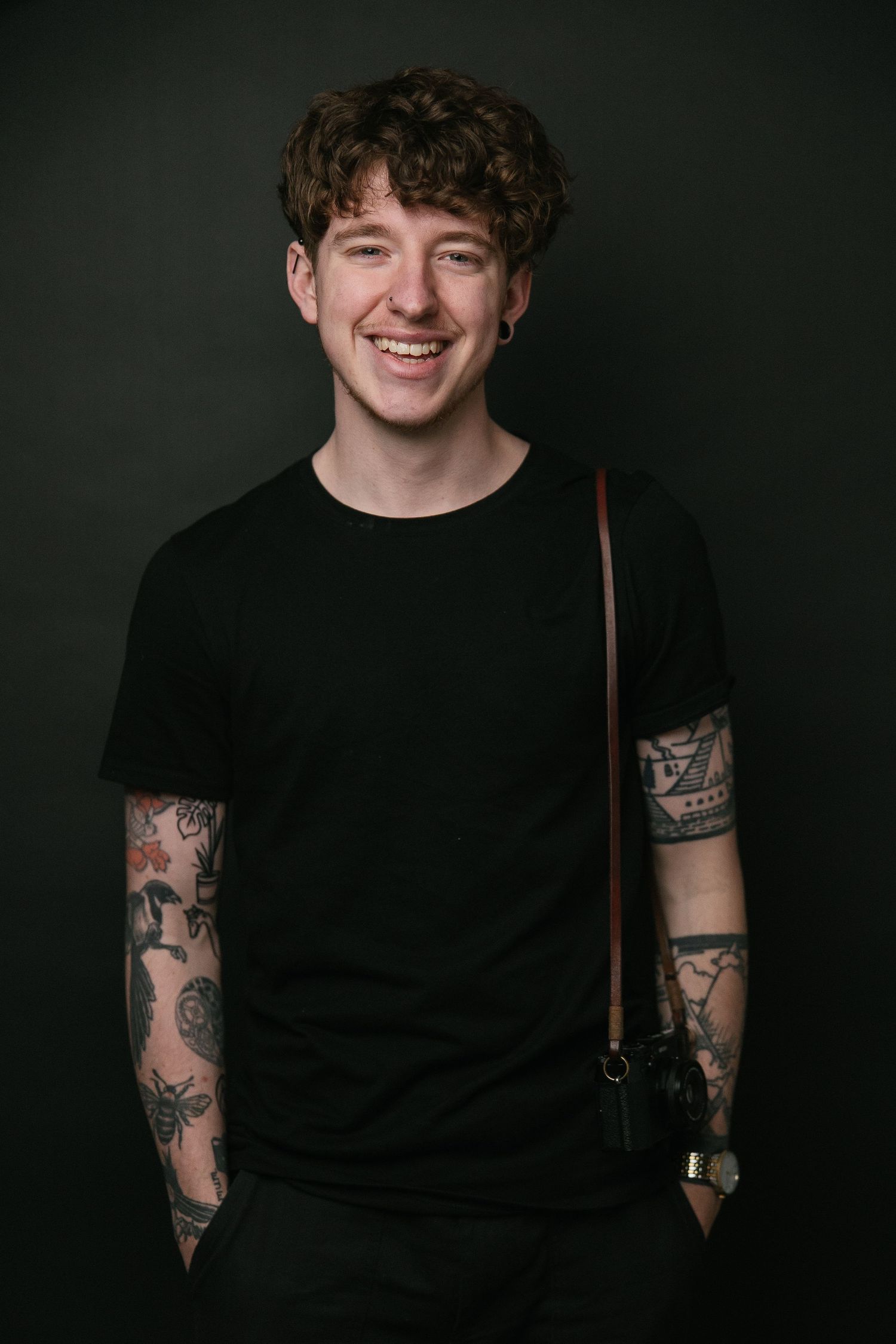 Young person in black t-shirt with sleeve tattoos smiling against dark background.