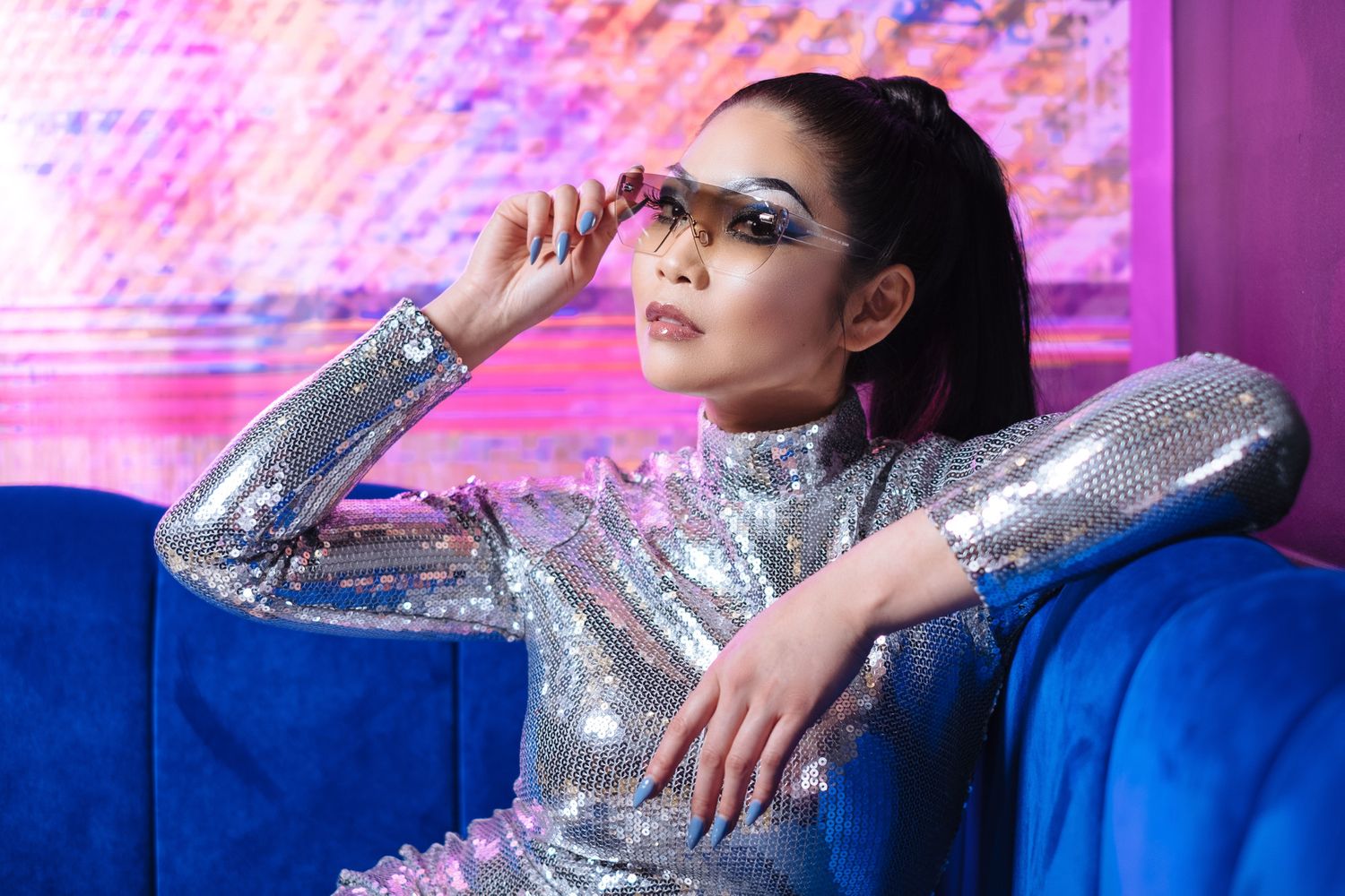 Glamorous model in silver sequined dress posing on blue couch with pink lighting.