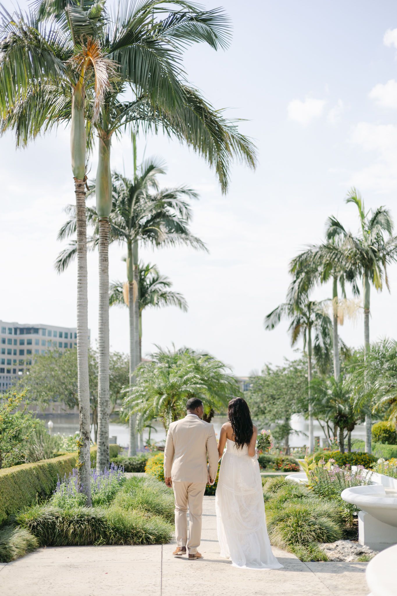 bride and groom holding hands on their wedding day as they walk through a garden between Bradenton palm trees.