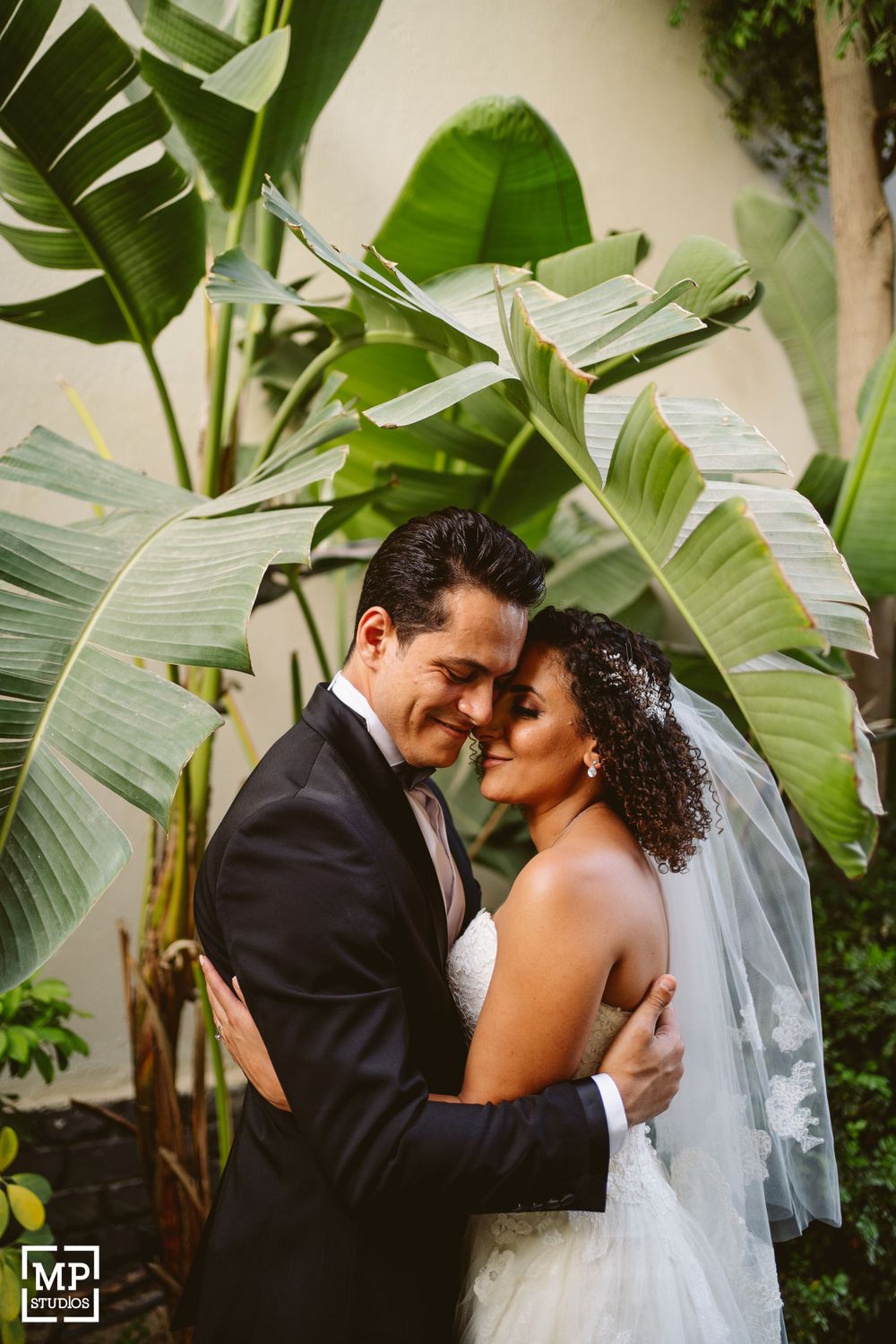 Newlyweds embrace tenderly under large tropical leaves during their wedding photoshoot.