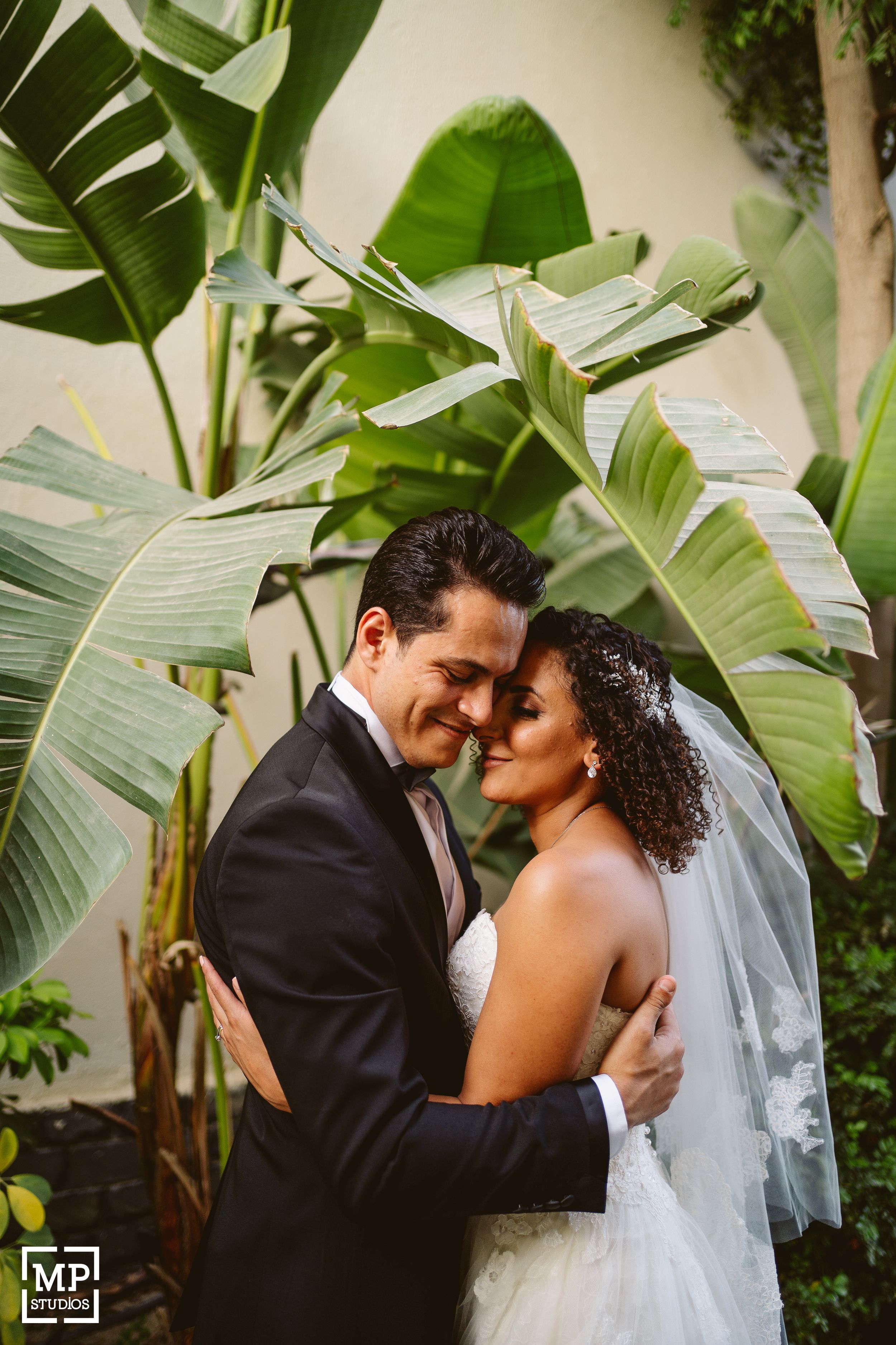 Newlyweds embrace tenderly under large tropical leaves during their wedding photoshoot.