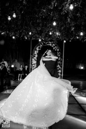 Dramatic black and white photo of wedding couple's first dance under twinkling lights and floral arch.