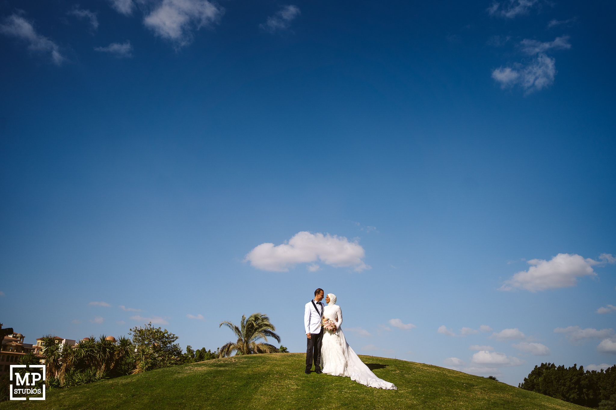 A bride and groom stand together on a hilltop against a bright blue sky with white clouds.