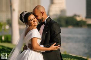 Elegant wedding couple embraces by waterfront with cityscape in background.