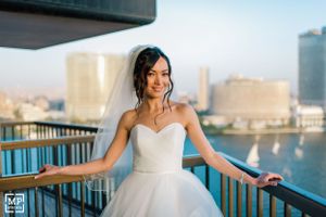 Bride in strapless gown poses joyfully on balcony with waterfront cityscape behind her.