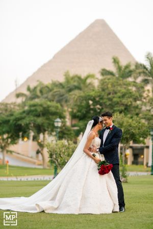 Wedding couple poses against backdrop of pyramid-shaped building and palm trees.