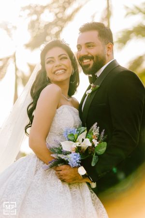 A joyful wedding couple smiles together while holding a white floral bouquet in outdoor sunlit setting.
