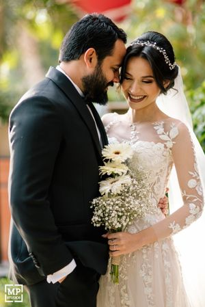 A romantic wedding moment with a bride in lace dress holding white flowers and groom in black tuxedo sharing an intimate moment.