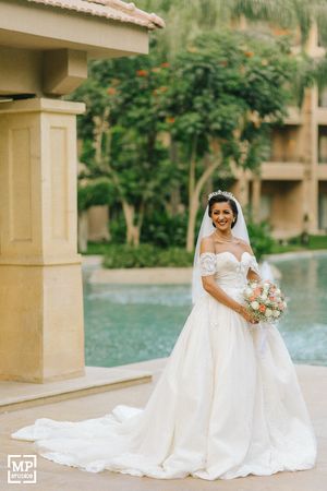 Bride in elegant off-shoulder gown poses by resort pool with bouquet.