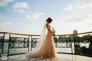 Bride in flowing tulle gown stands on balcony overlooking cityscape at sunset with bouquet.