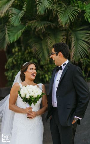 Wedding couple laughs together in front of tropical palm fronds while holding white bouquet.