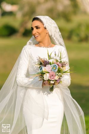 Bride in long sleeve lace gown smiles while holding pastel bouquet in outdoor garden setting.