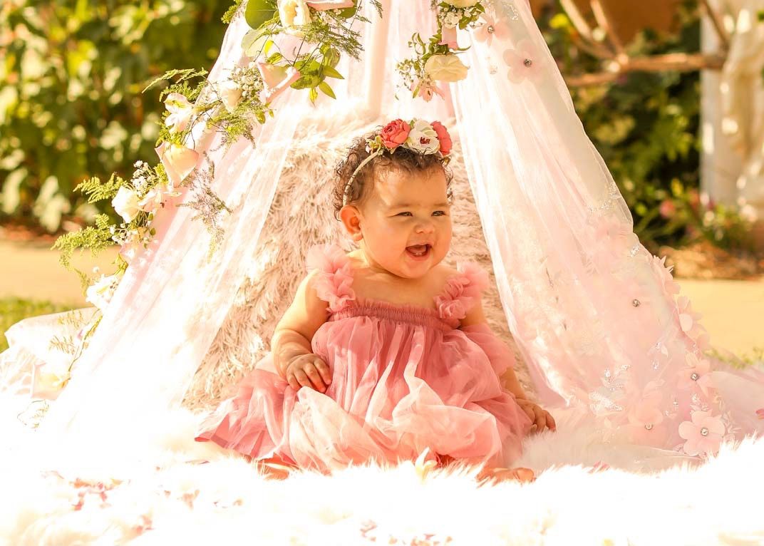 Baby in pink dress playing under delicate white lace tent in sunny garden setting.