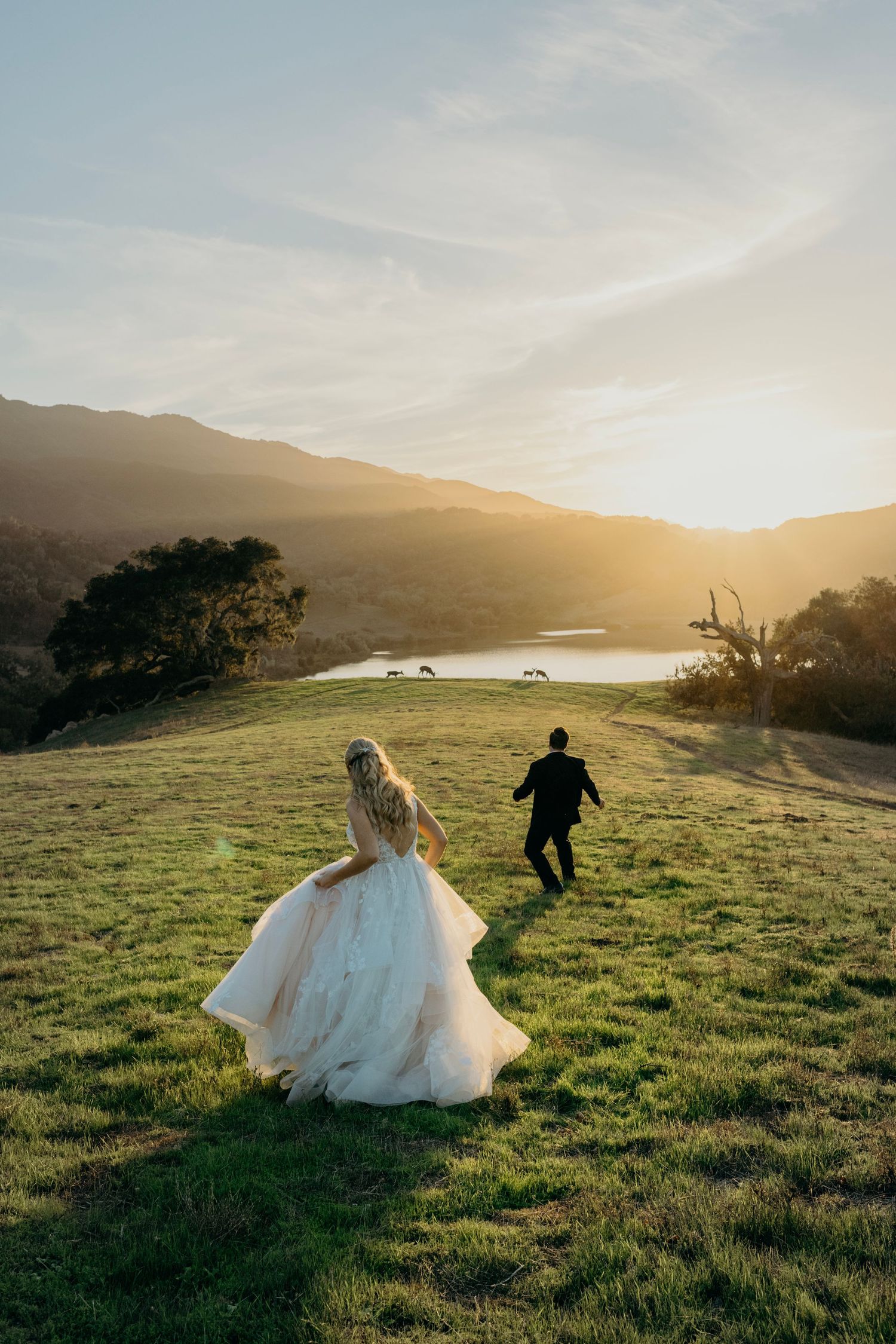 A couple runs playfully through a sunlit meadow during their wedding photoshoot at golden hour.