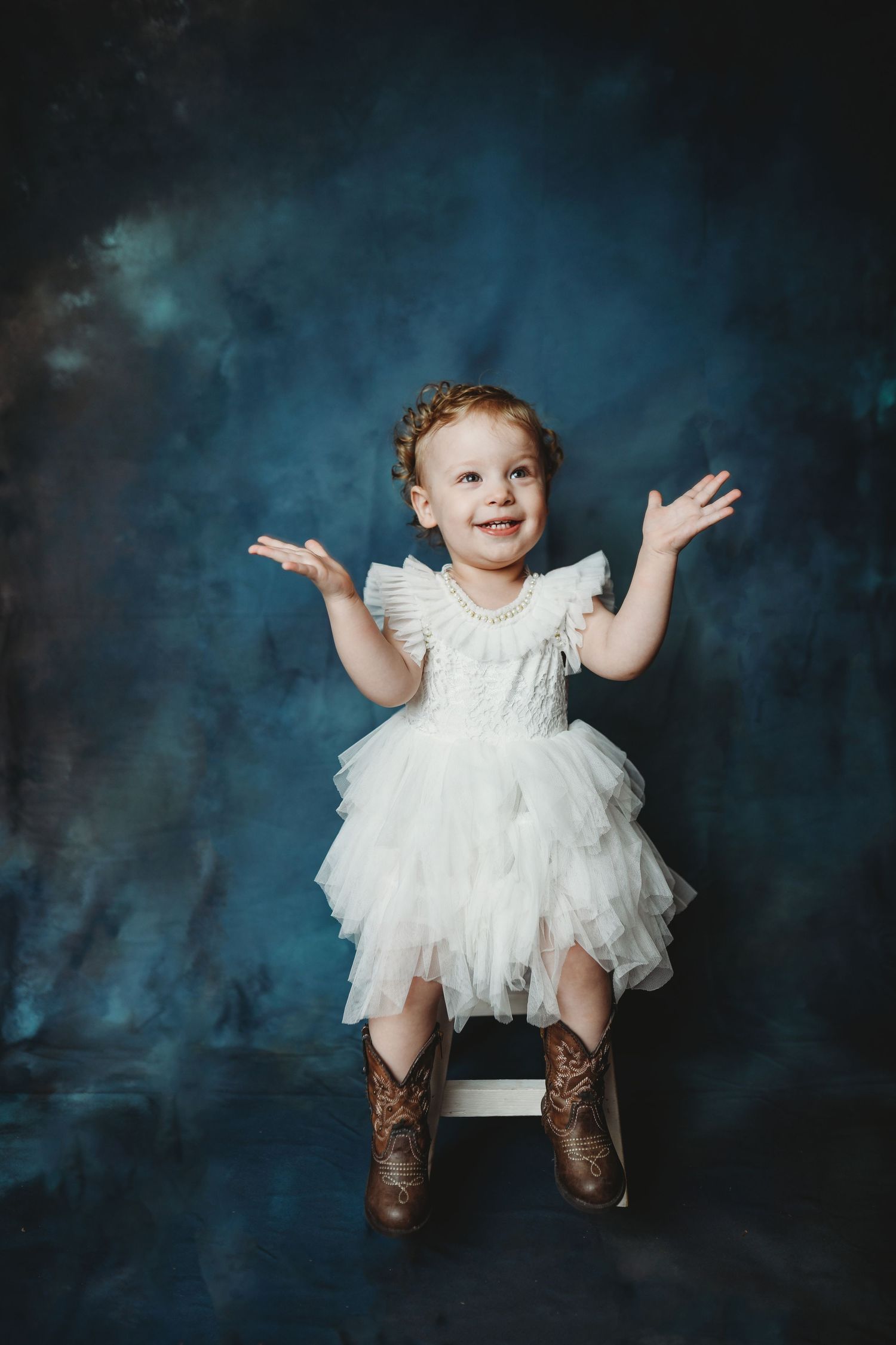 Small child in white dress and boots poses playfully against blue backdrop.