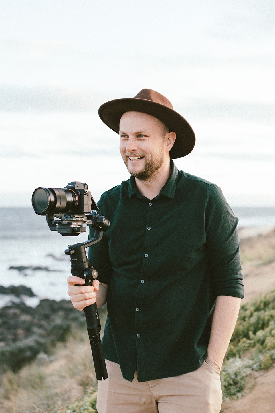 Photographer with camera gimbal equipment on coastal cliffside wearing dark hat and shirt.