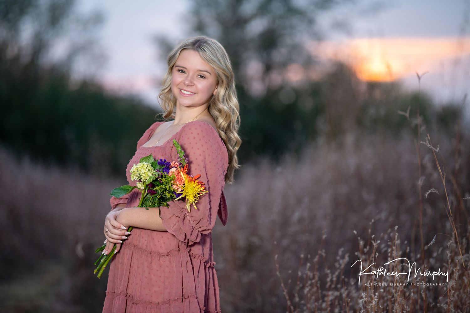Woman in dusty pink dress holds sunflower bouquet while posing in field during golden hour sunset.
