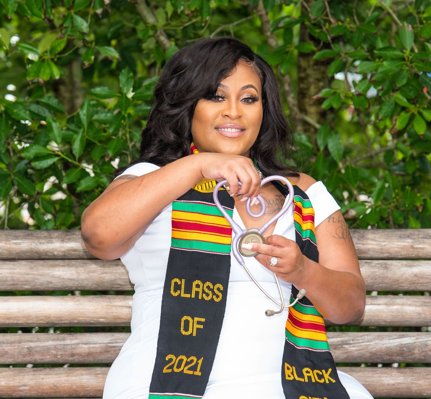Graduate in white dress with colorful graduation stole poses on wooden bench.