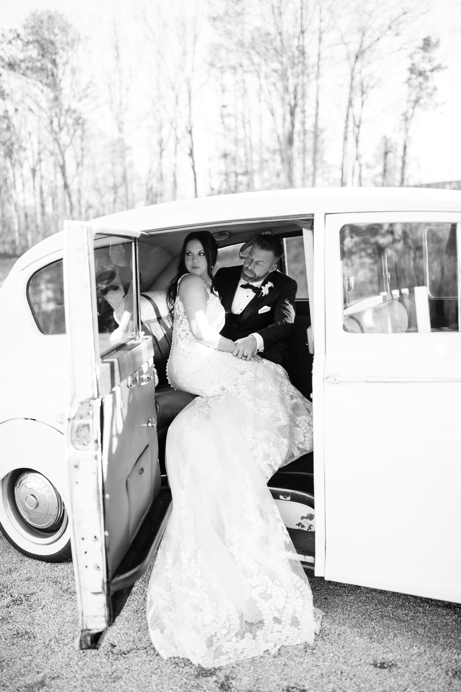 Wedding couple entering vintage white car with flowing lace dress trailing behind.