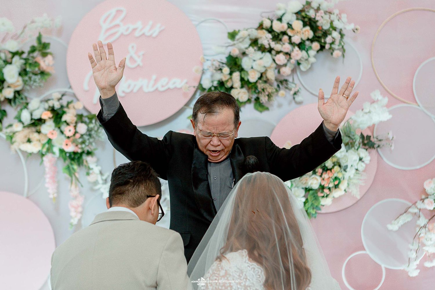 Wedding officiant raises hands during ceremony blessing against pink floral backdrop.
