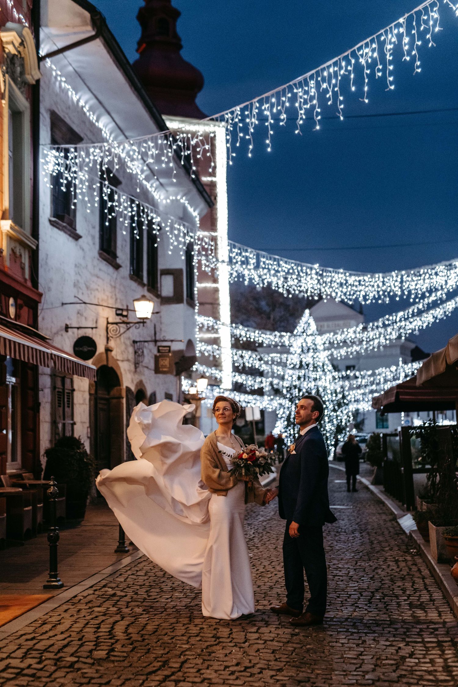 A wedding couple poses on cobblestone street under twinkling white lights at night.