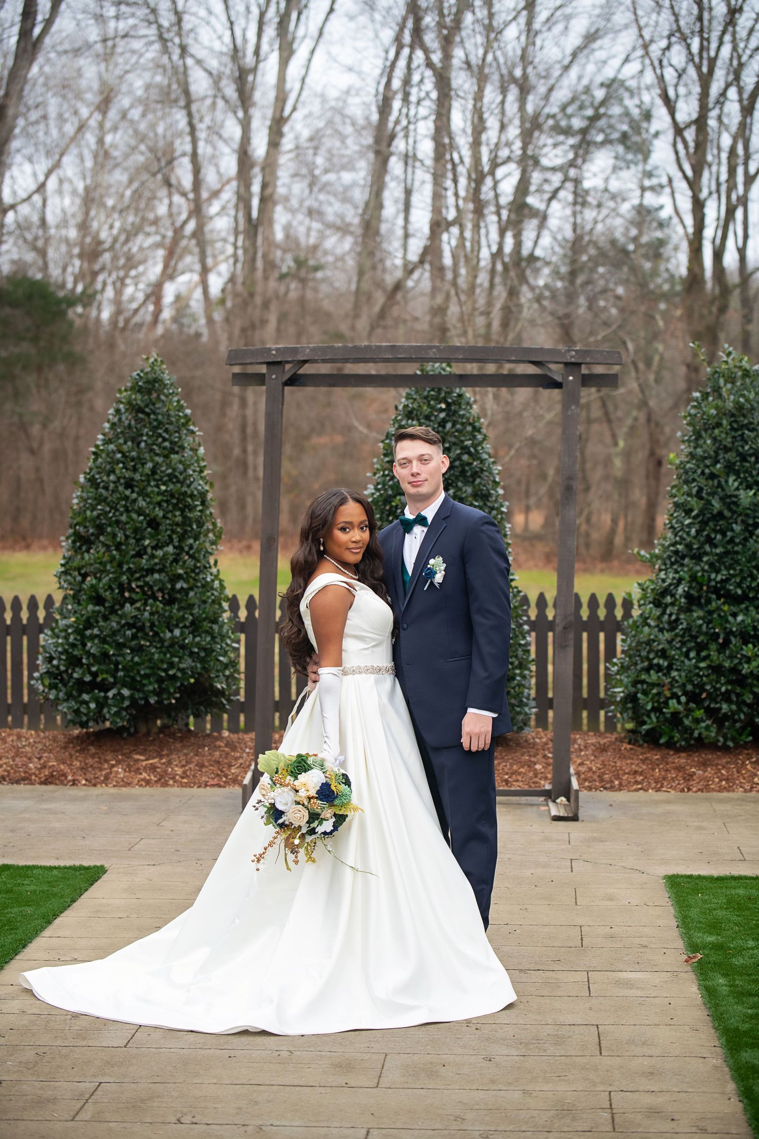 A wedding portrait of newlyweds posing in front of an outdoor arch and topiary trees.