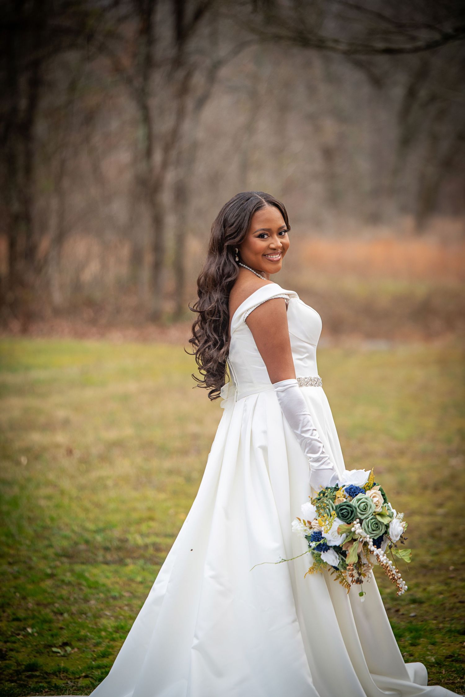 A radiant bride in a white gown and long gloves poses with her bouquet in a natural setting.