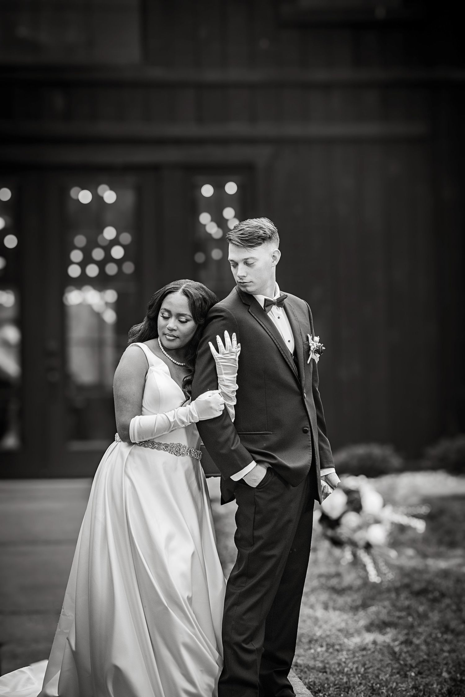 Couple in formal wedding attire share an intimate black and white moment with soft bokeh lighting in the background.