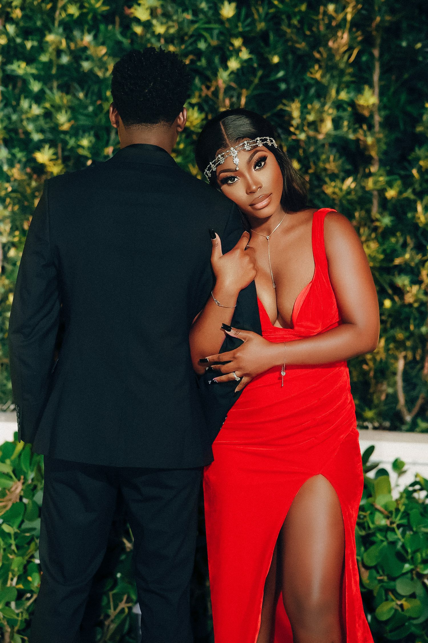 Woman in red gown with jeweled headpiece posing with man in black suit outdoors.