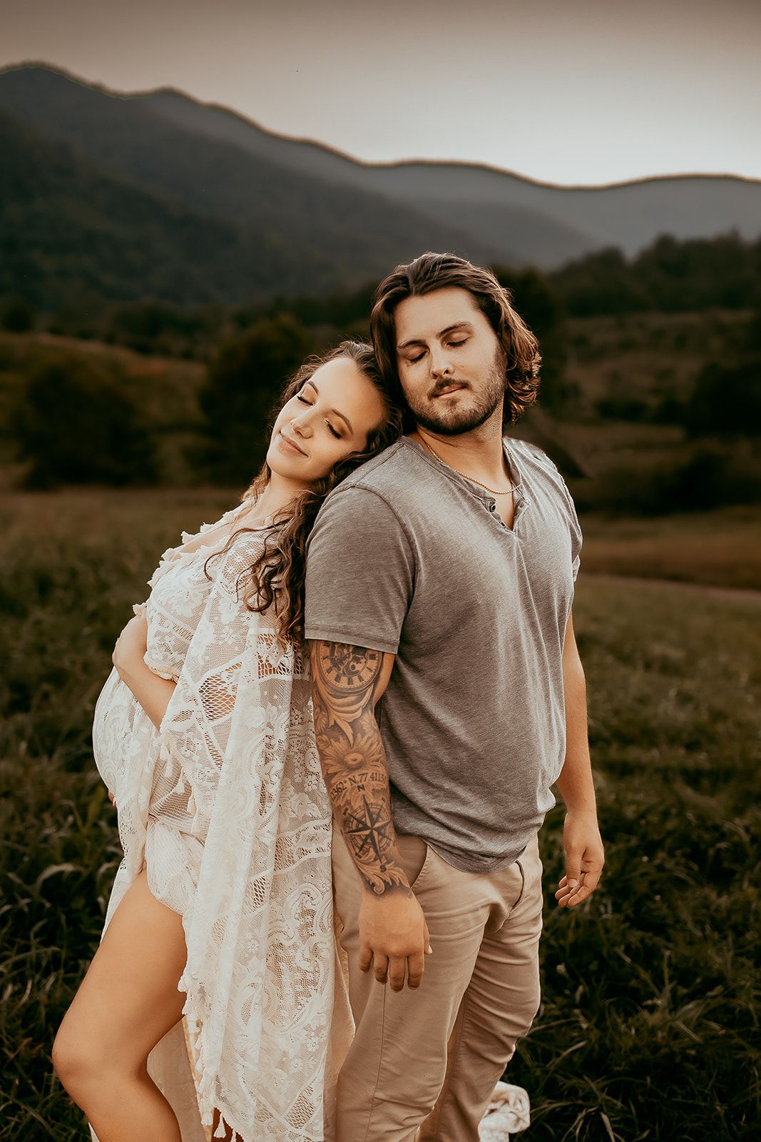 A woman stands back to back with her husband while holding her pregnant belly.