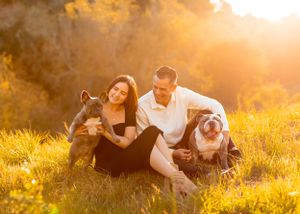A happy couple poses with their bulldogs in a sun-drenched field.