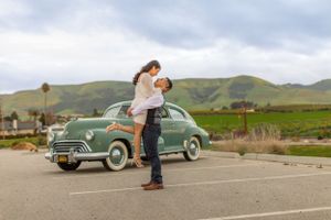 A romantic moment in front of a vintage green car with rolling hills in the background at sunset.