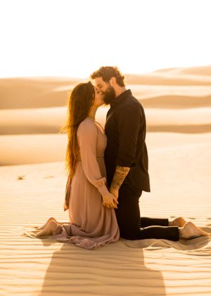 A romantic couple sits together on sand dunes during golden hour sunset.