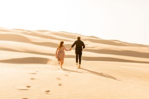 Couple holding hands while running across desert sand dunes at sunset.