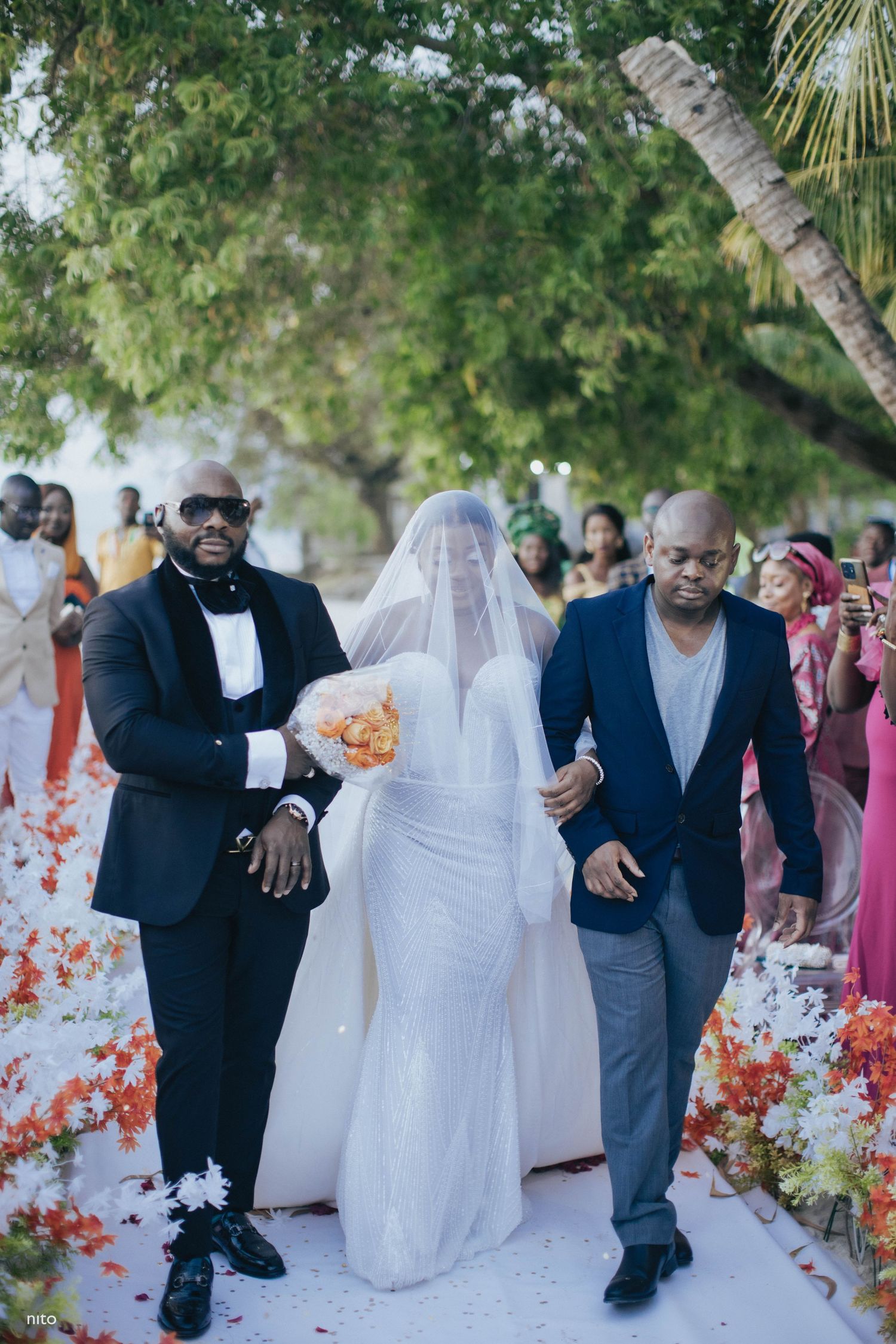 A wedding ceremony procession on a flower-lined path with attendants in formal attire.