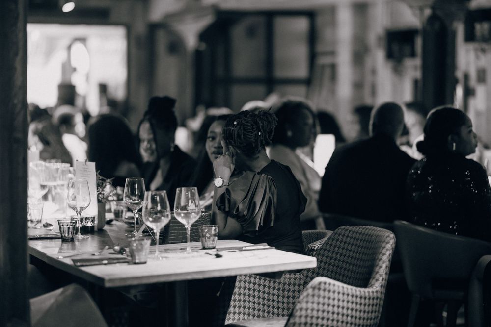 A black and white photograph of diners seated at tables with wine glasses in a dimly lit restaurant.