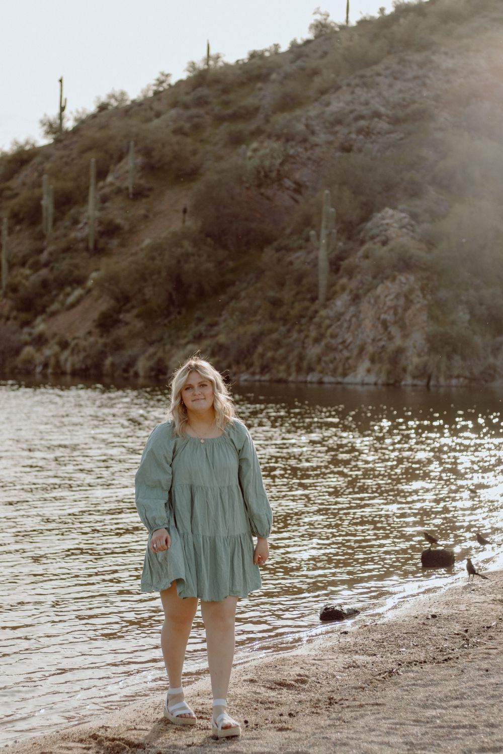 A tranquil scene at a desert lake with tall saguaro cacti on a hillside in the background.