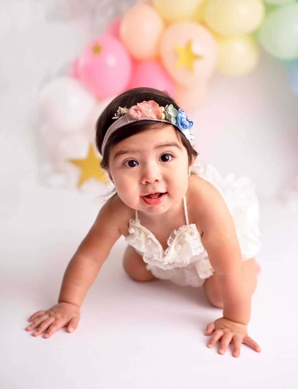 Baby in white lace dress crawls on white backdrop with pastel balloon arch and floral headband decoration.