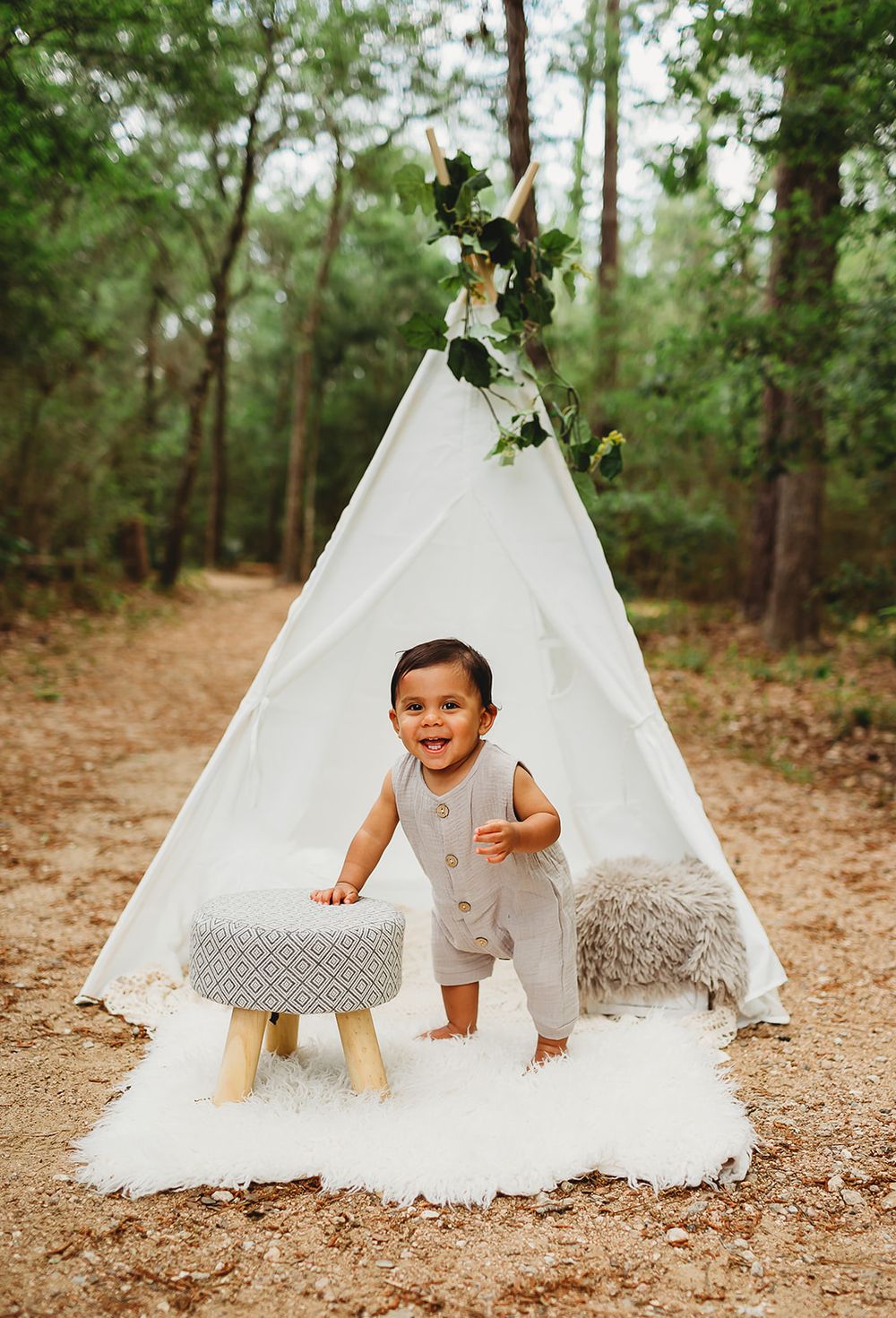 A white teepee tent decorated with greenery sits on a wooded path with a small stool and fuzzy cushions.