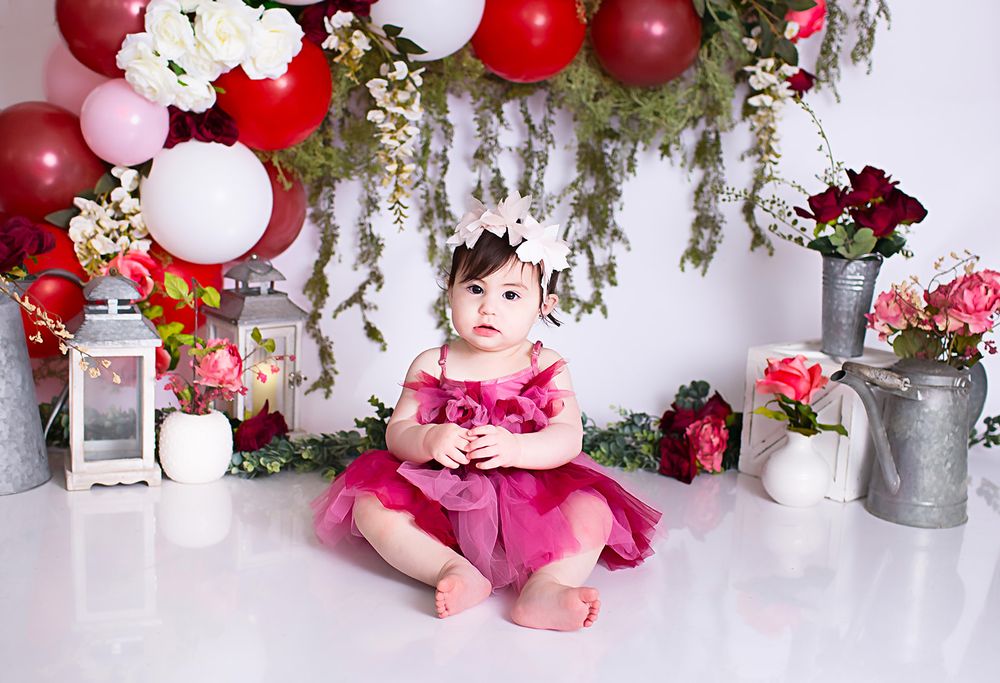 Festive setup with red and white balloon arch, lanterns, flowers and greenery decorations against white backdrop for first birthday.