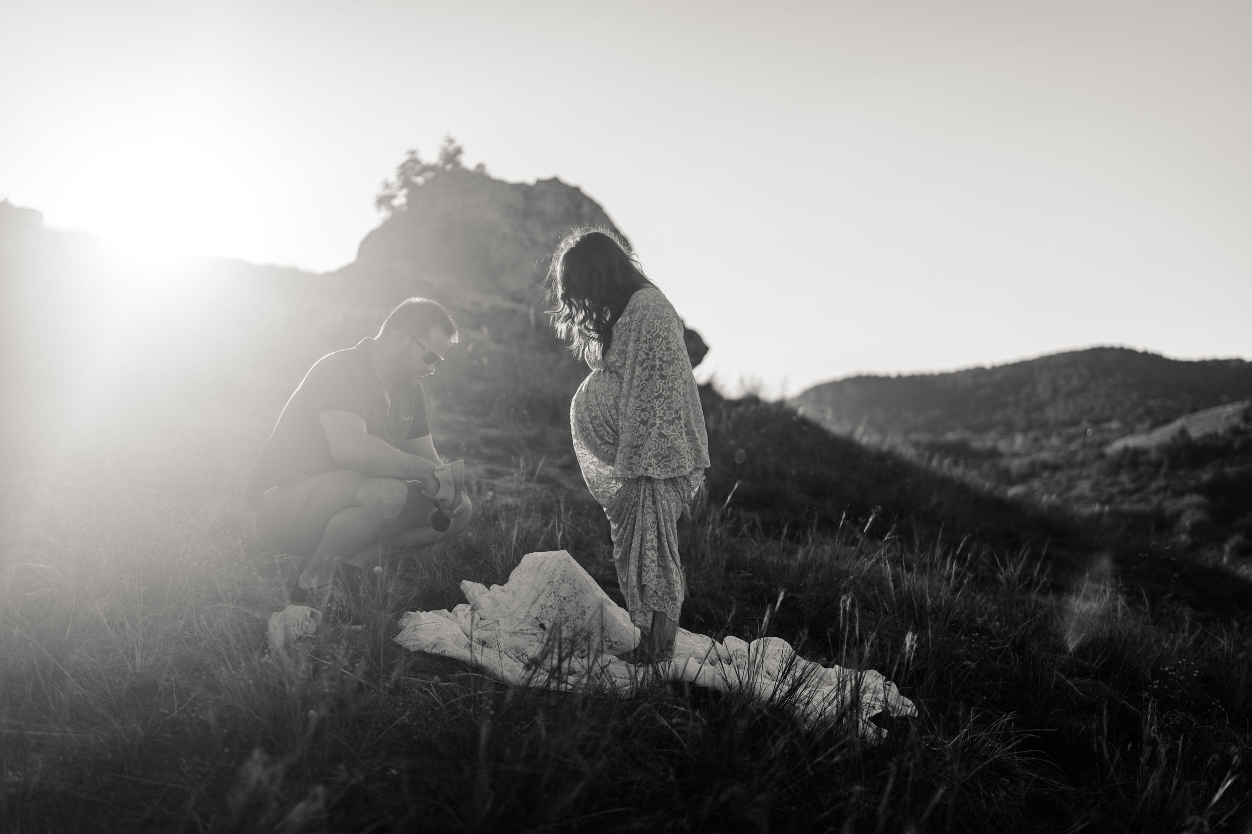 Silhouetted figure stands on rocky outcrop against bright sunlight during golden hour in black and white photography.