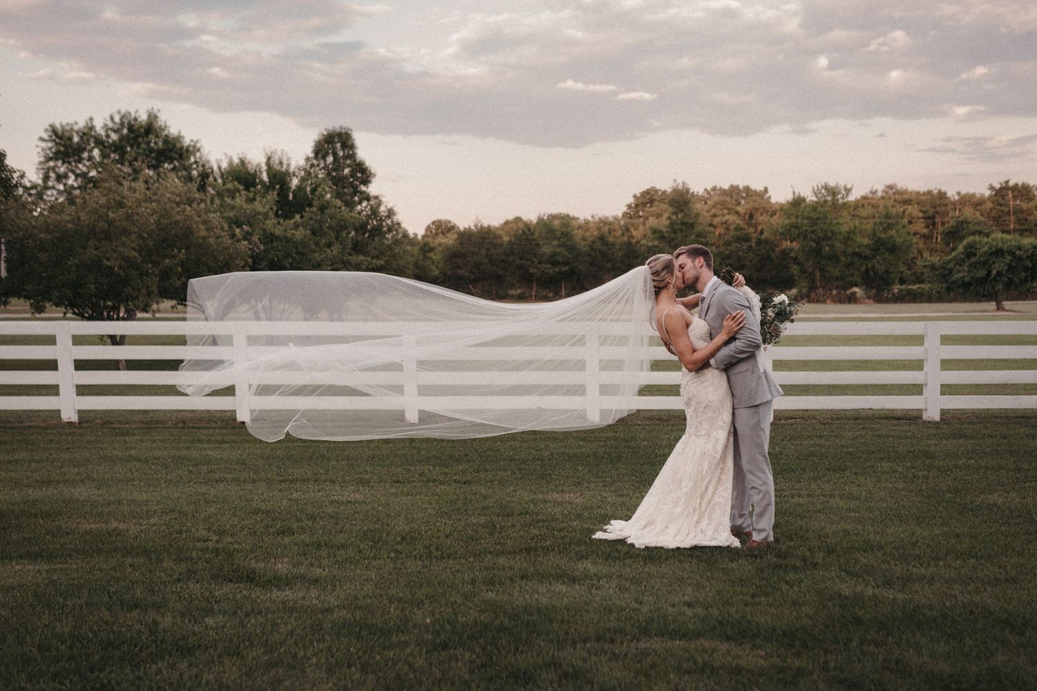 Newlyweds walk hand in hand across an open field with white fencing at sunset.