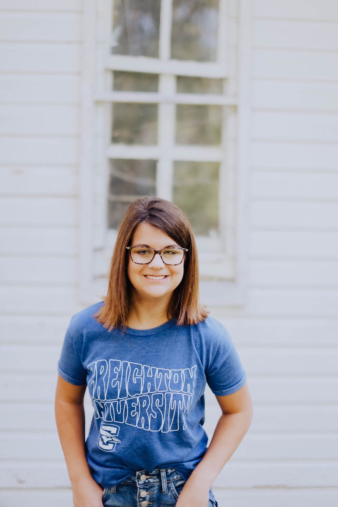 Person in glasses and blue collegiate t-shirt posing outdoors by white wall.