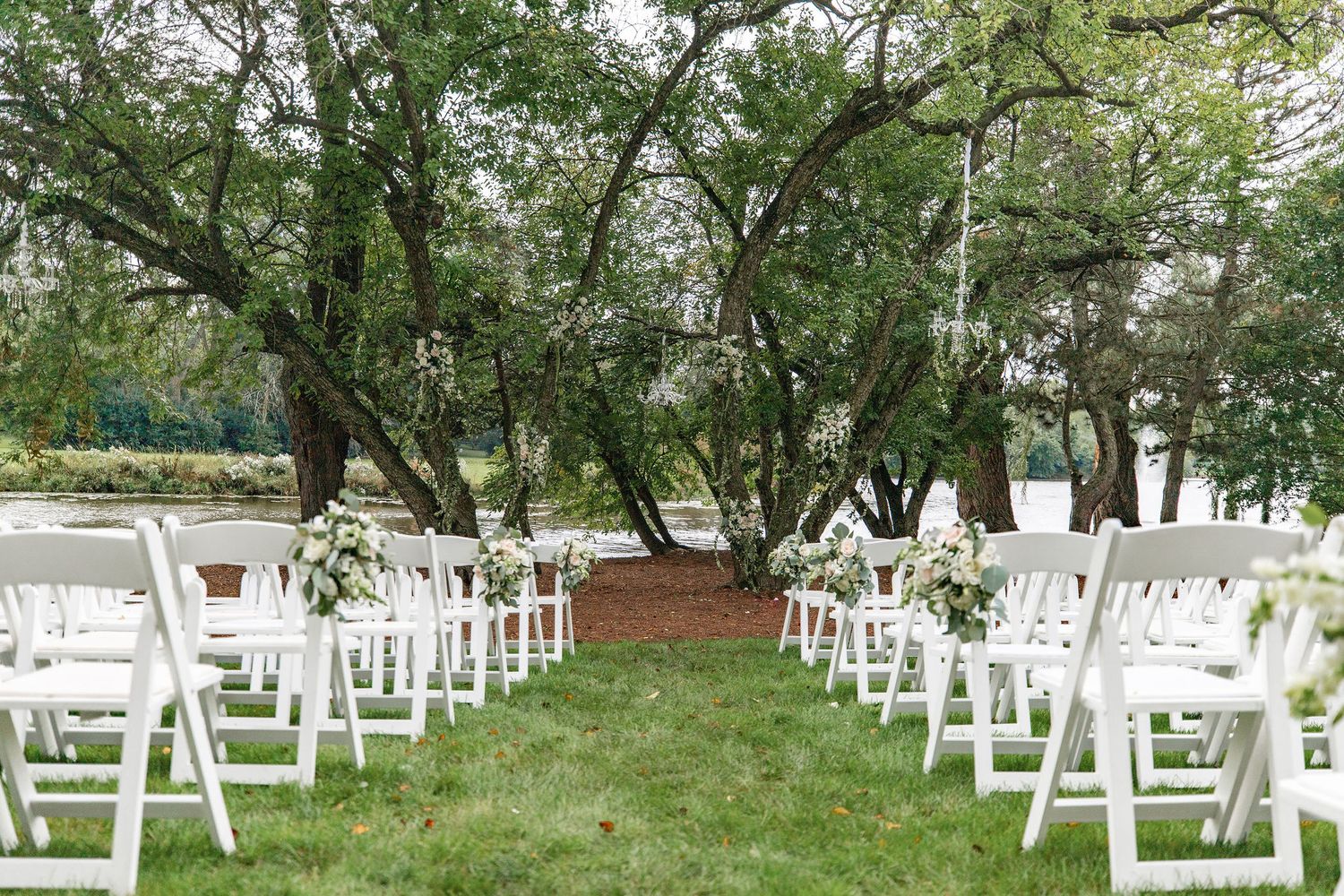White folding chairs arranged in rows for an outdoor wedding ceremony under shady trees.