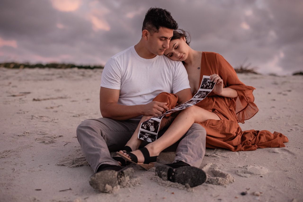 Couple sitting together on beach sand during sunset maternity photo session.