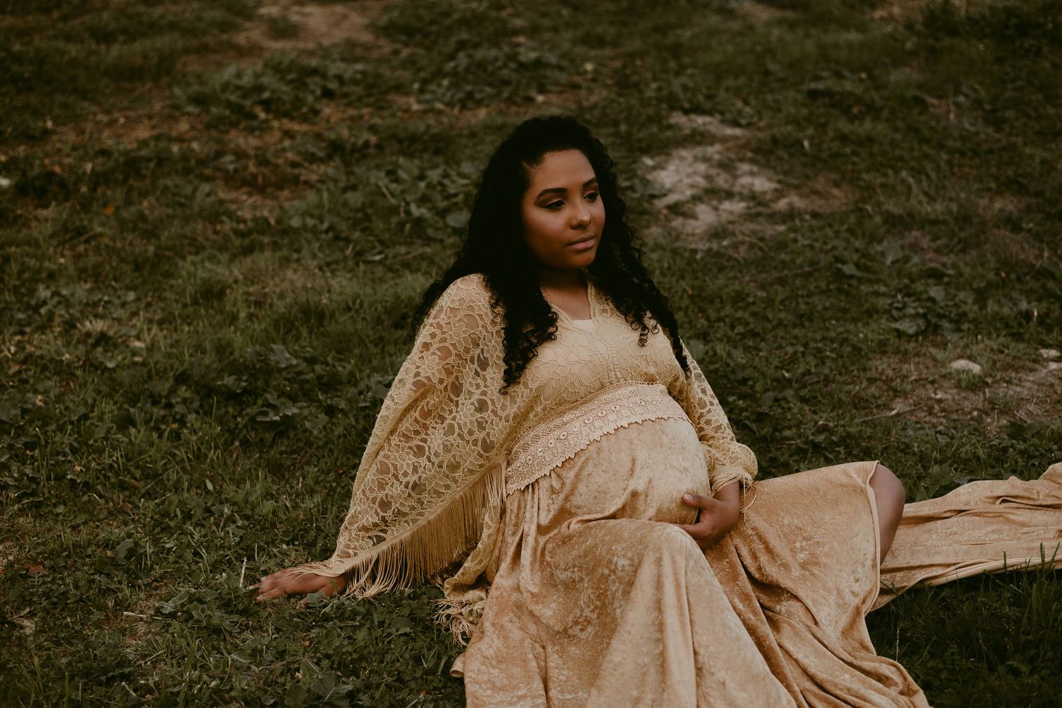 Expectant mother in flowing cream dress poses gracefully on grass in warm golden light.