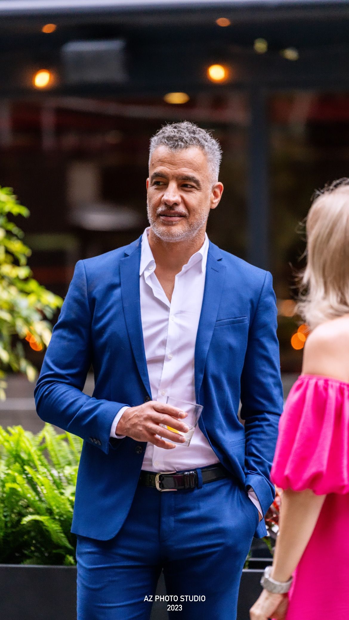 Person in blue suit holding drink at outdoor evening event.