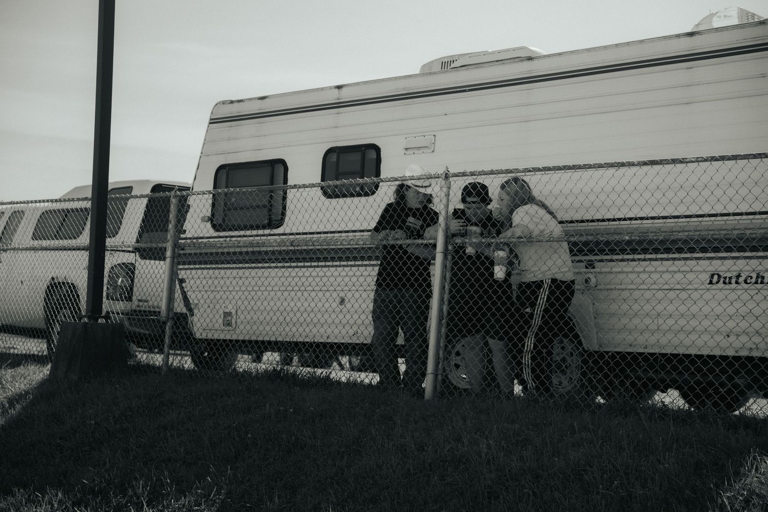 Vintage black and white photo of people gathered around RVs or trailers behind a chain link fence.